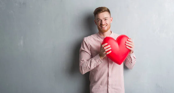 Young Redhead Man Grey Grunge Wall Holding Red Heart Happy — Stock Photo, Image