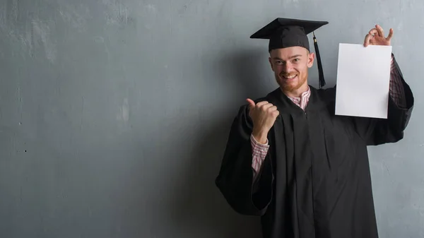 灰色のグランジの赤毛の若い男壁の身に着けている大学院制服指していると幸せそうな顔を笑顔で側まで親指で示すの学位を保持 — ストック写真