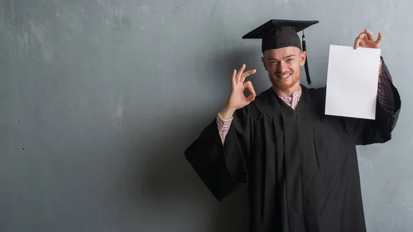 灰色のグランジの赤毛の若い男壁の身に着けている大学院制服優秀なシンボルの指で サインをして学位を保持 — ストック写真