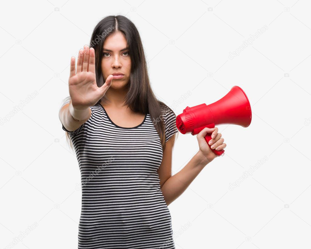 Young hispanic woman holding megaphone with open hand doing stop sign with serious and confident expression, defense gesture