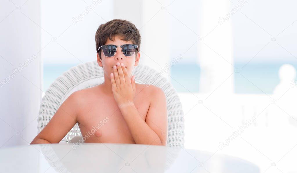 Young child on holidays sitting on a chair by the beach cover mouth with hand shocked with shame for mistake, expression of fear, scared in silence, secret concept