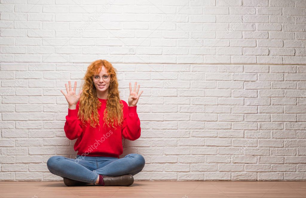 Young redhead woman sitting over brick wall showing and pointing up with fingers number nine while smiling confident and happy.
