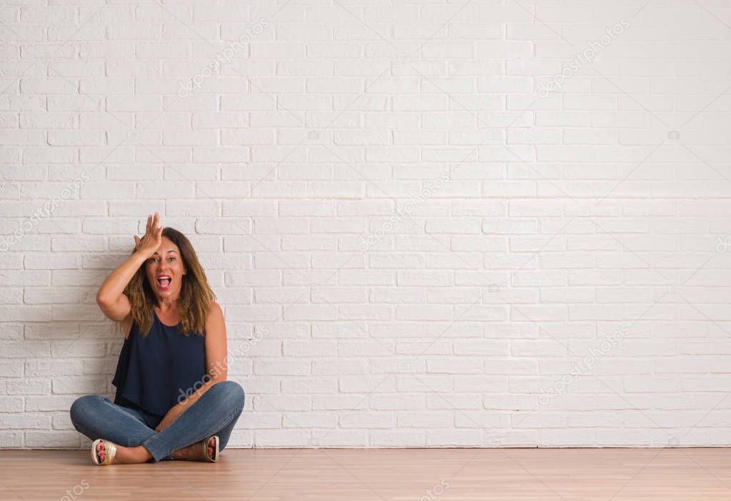 Middle age hispanic woman sitting on the floor over white brick wall surprised with hand on head for mistake, remember error. Forgot, bad memory concept.