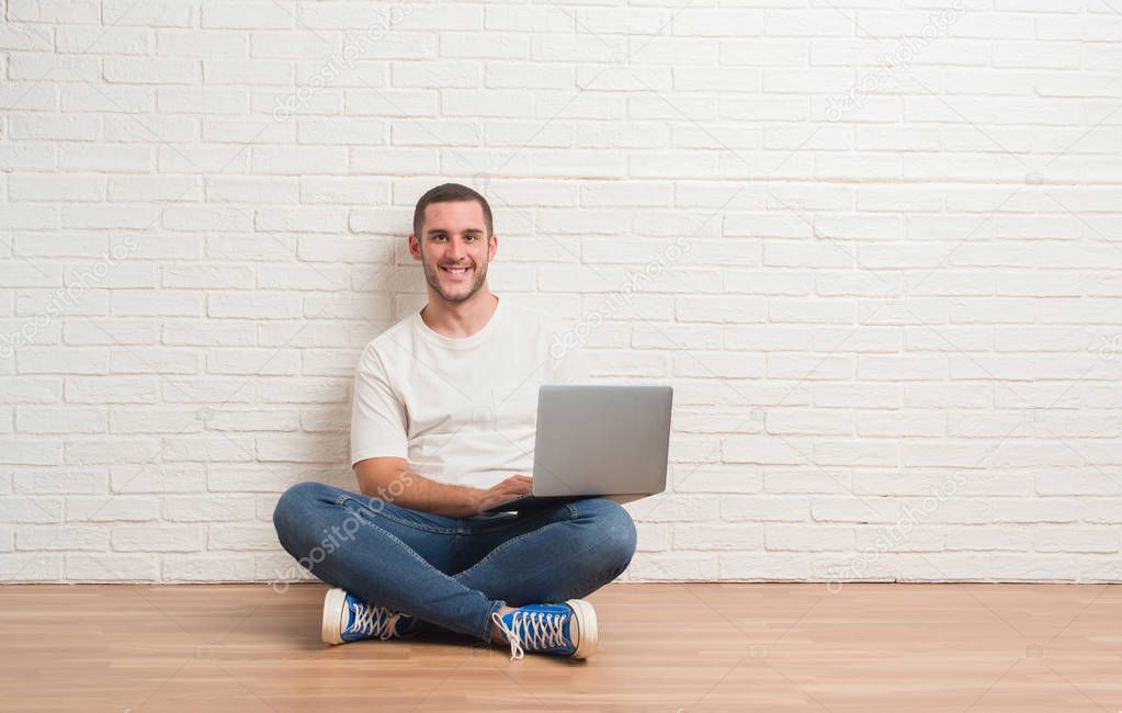 Young caucasian man sitting over white brick wall using computer laptop with a happy face standing and smiling with a confident smile showing teeth