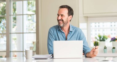 Orta yaş adam ayakta ve diş gösterilen kendine güvenen bir gülümsemeyle gülümseyerek mutlu bir yüz ile dizüstü evde kullanma