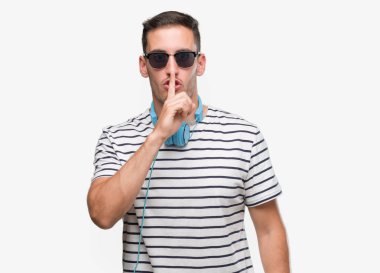 Handsome young man wearing headphones asking to be quiet with finger on lips. Silence and secret concept. clipart
