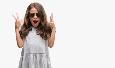 Brunette hispanic girl wearing sunglasses celebrating mad and crazy for success with arms raised and closed eyes screaming excited. Winner concept clipart