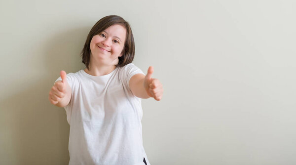 Down syndrome woman standing over wall approving doing positive gesture with hand, thumbs up smiling and happy for success. Looking at the camera, winner gesture.