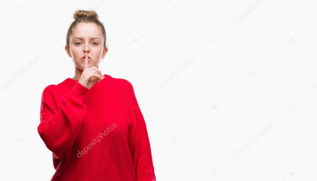 Young blonde woman wearing bun and red sweater asking to be quiet with finger on lips. Silence and secret concept.