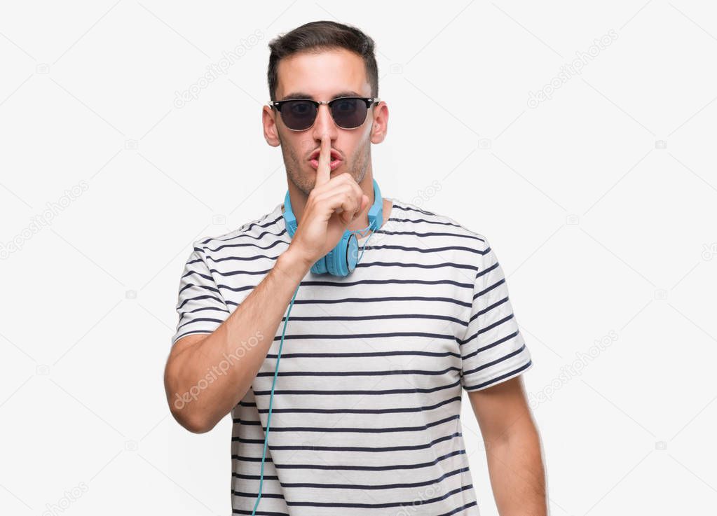 Handsome young man wearing headphones asking to be quiet with finger on lips. Silence and secret concept.