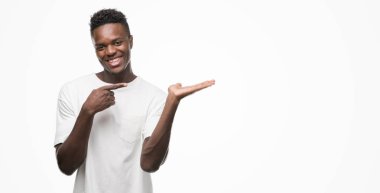 Hayret ve el ile sunulması ve parmak ile işaret ederken kameraya gülümseyen beyaz t-shirt giyen genç Afro-Amerikan adam.