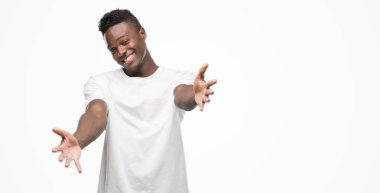 Sarılmak için kollarını açarak gülümseyerek kameraya bakarak beyaz t-shirt giyen genç Afro-Amerikan adam. Neşeli ifade kucaklayan mutluluk.