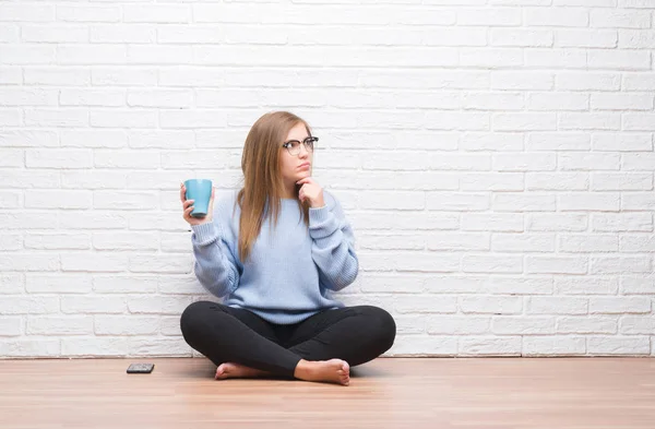 若い成人女性を白いレンガ壁煮沸質問 非常に混乱しているアイデアについて真面目な顔考え コーヒー飲みながら床に座って — ストック写真
