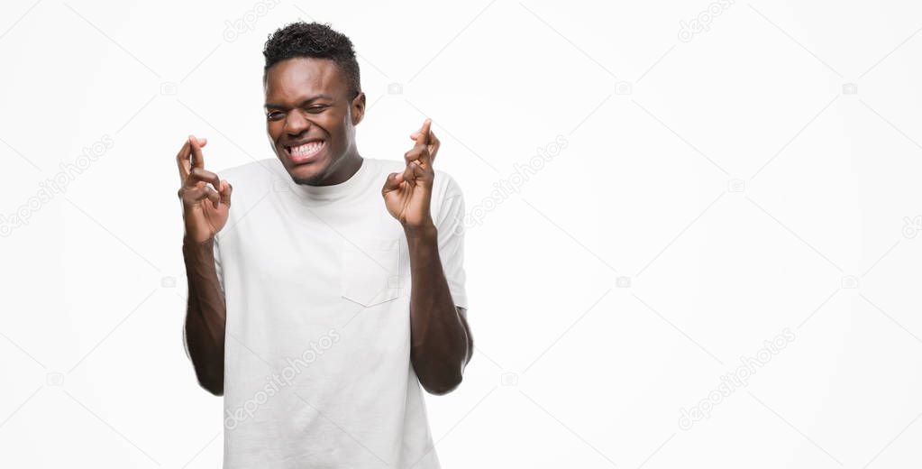 Young african american man wearing white t-shirt smiling crossing fingers with hope and eyes closed. Luck and superstitious concept.