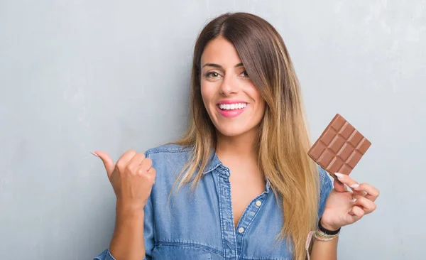 指していると幸せそうな顔を笑顔で側に親指を示すバー チョコレートを食べる灰色グランジ壁を越えて若い成人女性 — ストック写真