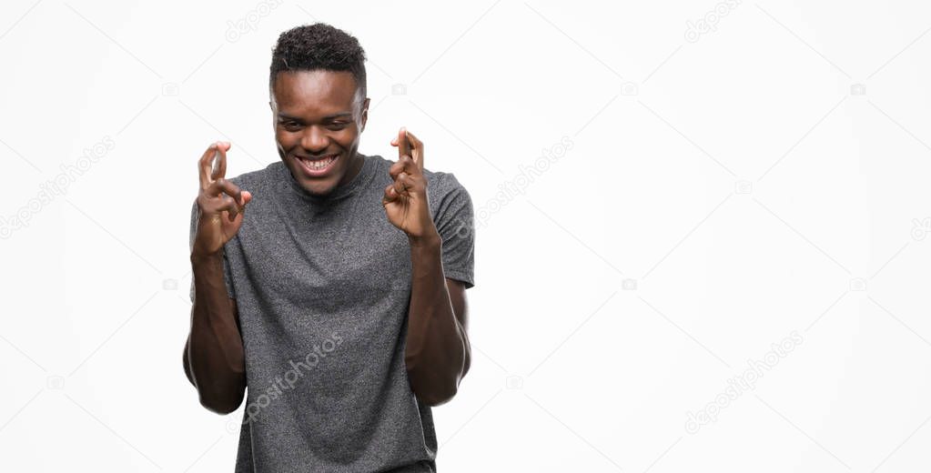 Young african american man wearing grey t-shirt smiling crossing fingers with hope and eyes closed. Luck and superstitious concept.