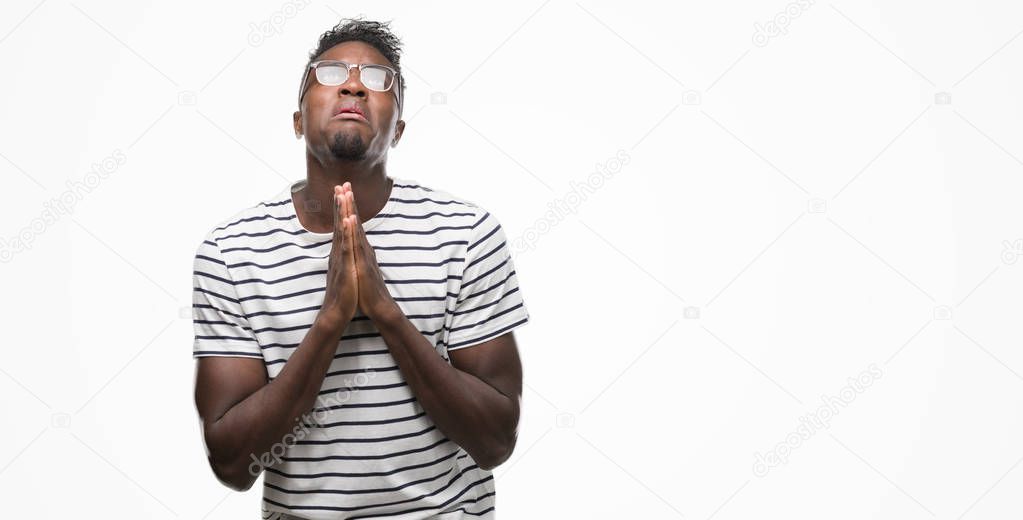 Young african american man wearing glasses and navy t-shirt begging and praying with hands together with hope expression on face very emotional and worried. Asking for forgiveness. Religion concept.