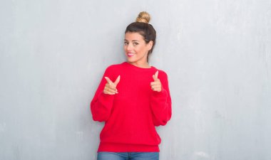 Gri grunge duvar kış kıyafeti işaret parmakları için fotoğraf makinesi ile mutlu ve komik yüz giyen genç yetişkin kadın. İyi enerji ve vibes.