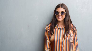 Grunge gri duvar ayakta ve diş gösterilen kendine güvenen bir gülümsemeyle gülümseyerek mutlu bir yüz ile retro güneş gözlüğü ve kıyafet giyen genç esmer kadın