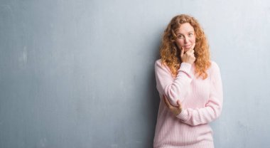 Genç redhead gri grunge duvar giyen üzerinde pembe kadın kazak emin çapraz kollar ve çeneme kaldırdı el ile gülümseme ile kameraya bakıyor. Pozitif düşünce.
