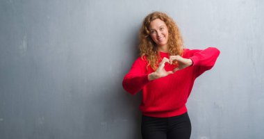 Genç redhead gri grunge duvar giyen üzerinde kırmızı kadın kazak aşık kalp simgesi ve elleriyle şekil gösteren gülümseyen. Romantik kavramı.