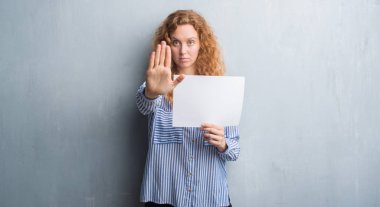 Gri grunge duvar holding boş kağıt sayfası açık eliyle dur işareti ile ciddi ve kendine güvenen ifade, savunma hareketi yaparken genç Kızıl saçlı kadın