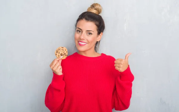 食べるチョコレート チップ クッキー ポイントと幸せそうな顔を笑顔で側に親指を示すグレー グランジ壁を越えて若い成人女性 — ストック写真