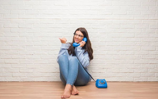 Young brunette woman sitting on the floor calling on vintage telephone pointing and showing with thumb up to the side with happy face smiling