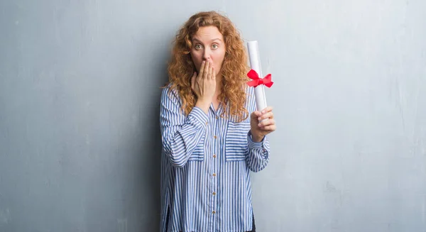 Young redhead business woman over grey grunge wall holding diploma cover mouth with hand shocked with shame for mistake, expression of fear, scared in silence, secret concept