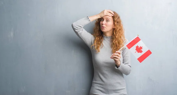 Young redhead woman over grey grunge wall holding flag of Canada stressed with hand on head, shocked with shame and surprise face, angry and frustrated. Fear and upset for mistake.