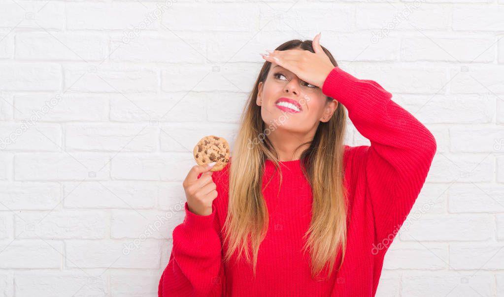 Young adult woman over white brick wall eating chocolate chip cooky stressed with hand on head, shocked with shame and surprise face, angry and frustrated. Fear and upset for mistake.