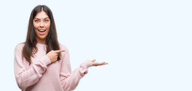 Hayret ve el ile sunulması ve parmak ile işaret ederken kameraya gülümseyen bir kazak giyen genç güzel İspanyol kadın.