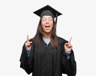 Mezun kap ve üniforma giyen genç İspanyol kadın hayrete ve yukarıya bakıyor ve parmakları ile işaret ve silah kaldırdı şaşırttı.