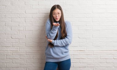 Genç Chinise kadın bir öpücük el ile güzel ve seksi olmak hava üfleme kamera bakıyor beyaz tuğla duvarın üzerinden. Aşk ifade.