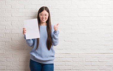 Boş kağıt ok işareti, başparmak ile parmak, mükemmel işareti yapıyor kocaman bir gülümseme ile mutlu tutarak tuğla duvarın üzerinden Genç Çinli kadın