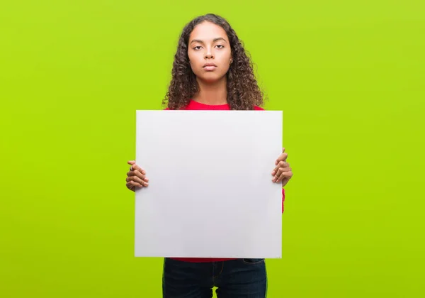 Young hispanic woman holding blank banner with a confident expression on smart face thinking serious