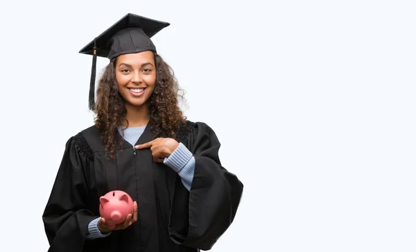 年轻的西班牙裔妇女穿着毕业制服持有小猪银行惊讶的脸指向自己的手指 — 图库照片