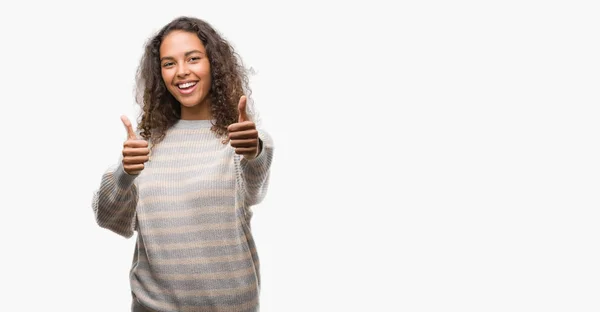 ストライプ セーターを承認する身に着けている美しい若いヒスパニック系女性は 笑顔と幸せな成功のため親指の手で肯定的なジェスチャーをしています カメラ 勝者のジェスチャーを見てください — ストック写真