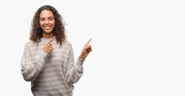笑顔で側に つの手と指で指しているカメラ目線のストライプ セーターを着ている美しい若いヒスパニック系女性 — ストック写真