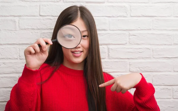 年轻的中国妇女在砖墙看通过放大镜与惊奇面孔指向自己手指 — 图库照片