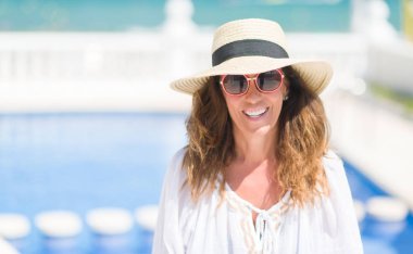 Orta yaş esmer kadın ayakta ve diş gösterilen kendine güvenen bir gülümsemeyle gülümseyerek mutlu bir yüz ile yüzme havuz