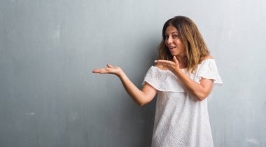 Orta yaş İspanyol kadın hayrete ve el ile sunulması ve parmak ile işaret ederken kameraya gülümseyen gri grunge duvarın üzerinden ayakta.