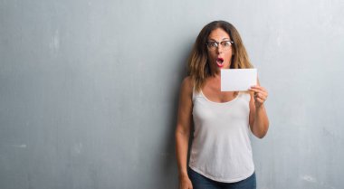 Gri grunge duvar holding boş kartı bir sürpriz yüz, korkuyor ve heyecanlı korku ifadesi ile şok korkmuş üzerinde duran orta yaş İspanyol kadın