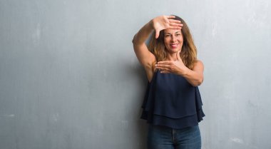 Elle yapma çerçeve ve parmakları ile mutlu yüz gülümseyen gri grunge duvarın üzerinden duran orta yaş İspanyol kadın. Yaratıcılık ve Fotoğrafçılık kavramı.