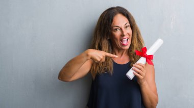 Diploma çok mutlu el ve parmak ile işaret tutan gri grunge duvarın üzerinden duran orta yaş İspanyol kadın