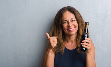 Orta yaş İspanyol kadın işaret ve başparmak kadar mutlu yüz gülümseyen tarafı ile gösterilen bira şişesi holding gri grunge duvarın üzerinden ayakta