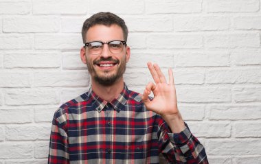 Genç yetişkin adam parmak, mükemmel sembolü ile ok işareti yapıyor beyaz tuğla duvarın üzerinden ayakta