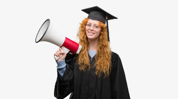 年轻红发学生女子穿着毕业制服持有扩音器与一个幸福的脸站在微笑着自信的微笑显示牙齿 — 图库照片