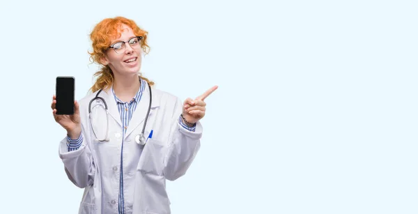 年轻的红发医生的女人显示智能手机非常高兴指向与手和手指的一侧 — 图库照片