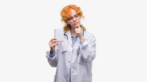 Jovem Ruiva Médico Mulher Segurando Notebook Branco Rosto Sério Pensando — Fotografia de Stock